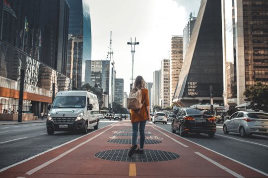 City street, with a woman in the center of the street walking away from the camera, wearing a red coat and tan backpack.